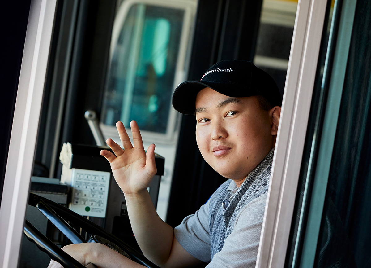 Closeup of bus operator looking to his left waving at the camera.